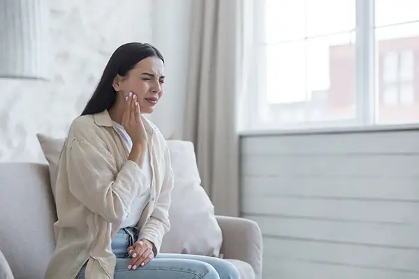 Young woman sitting on a couch, holding her cheek and wincing due to severe toothache, requiring emergency dental care.
